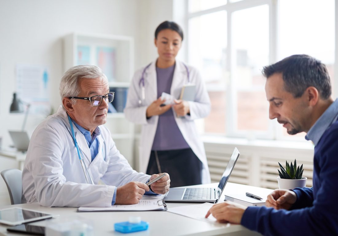 A doctor in a white coat talking across a desk with a patient, a second clinician standing behind them holding a notebook