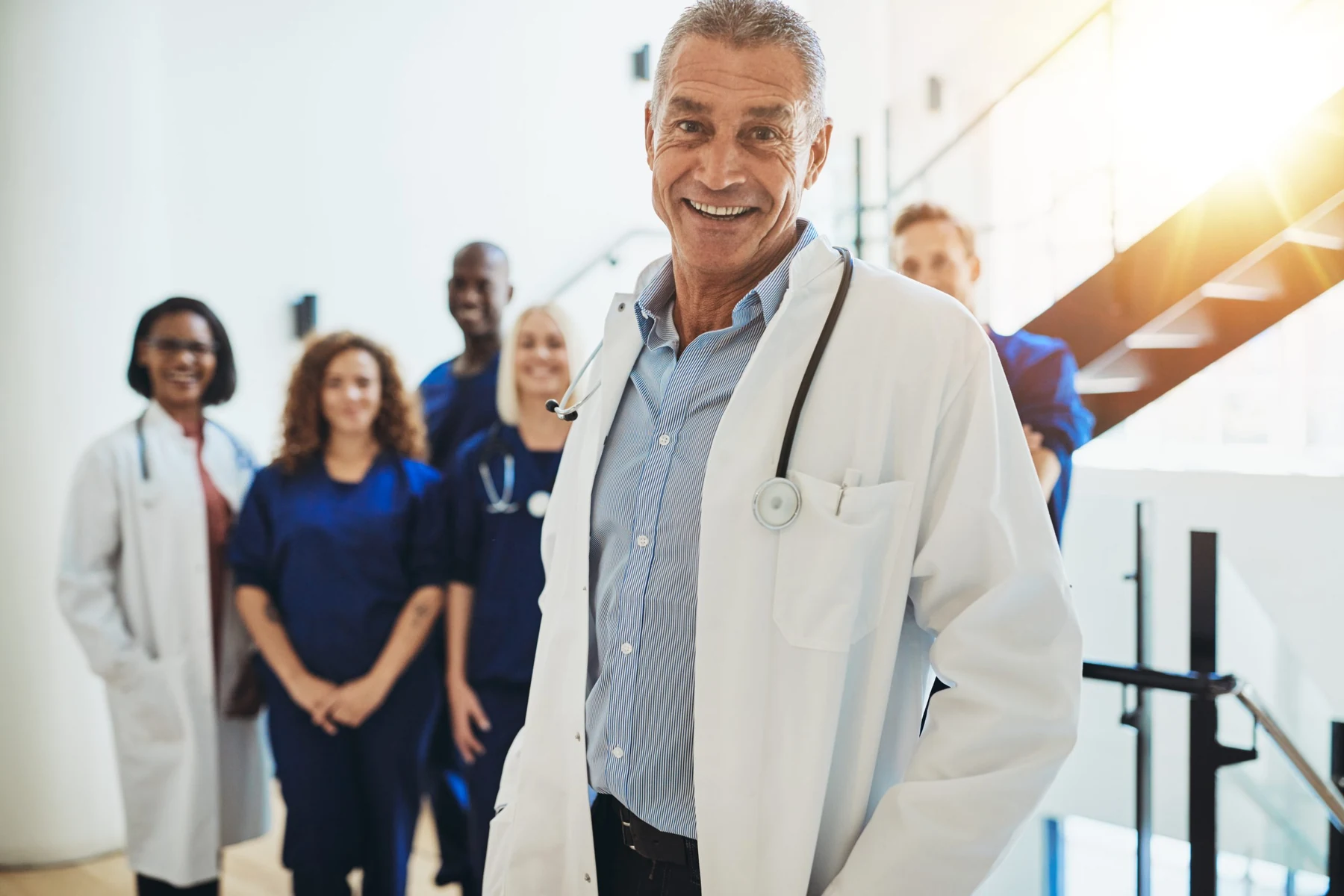 A smiling doctor in a white coat and stethoscope in a bright hospital stairwell, five colleagues in white coats and scrubs standing behind him