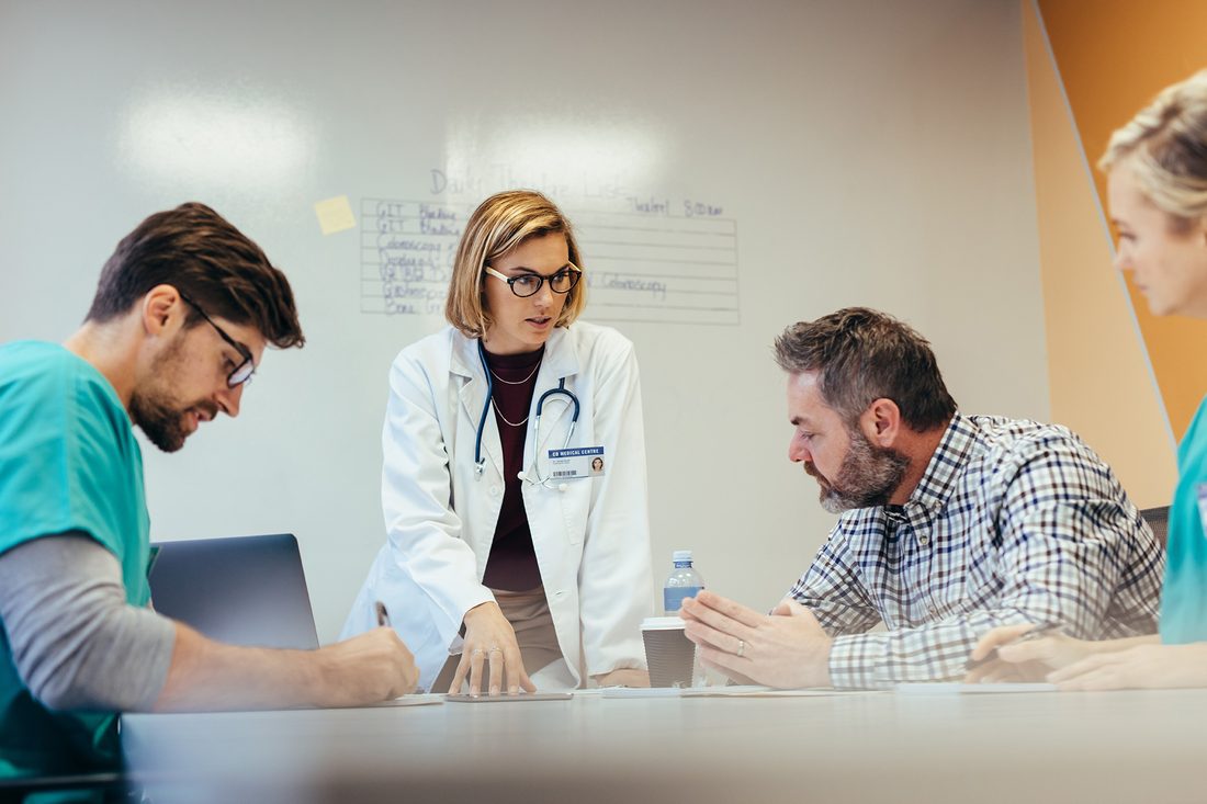 A doctor leaning over a meeting table talking with a colleague in scrubs and a visitor, a ward list on the board behind them