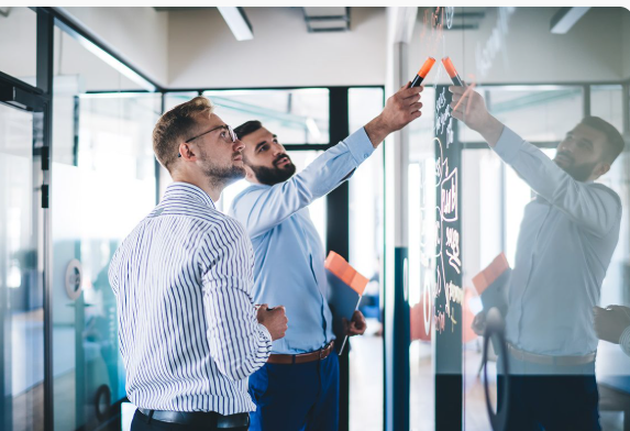 Two colleagues working through a plan in orange marker on a glass wall in an open office