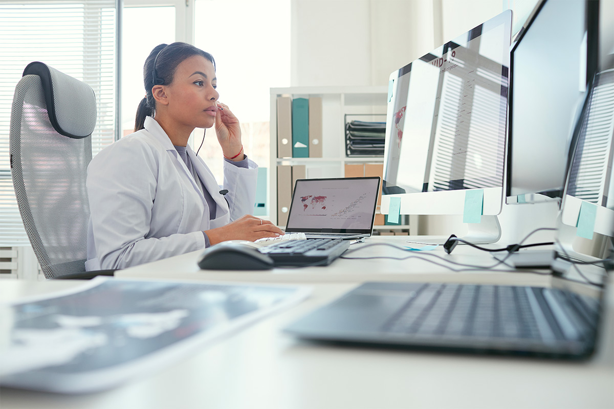 A clinician in a white coat and headset sits at a desk of monitors, typing on a laptop that shows a data dashboard, with a printed scan beside her keyboard.