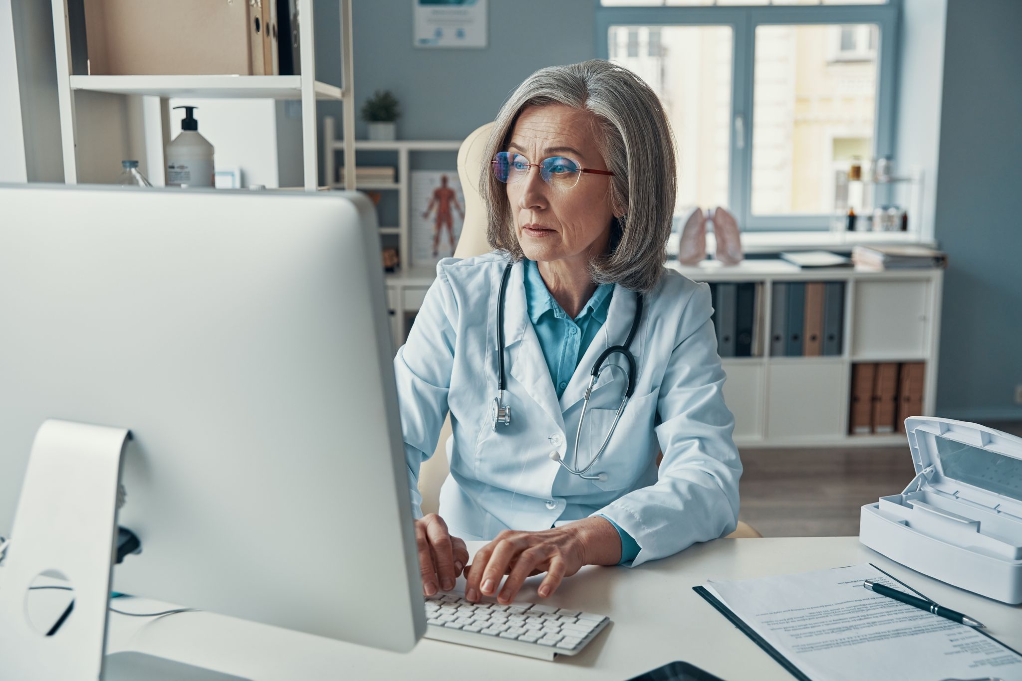 A physician working at a desktop computer in a consulting room, the screen turned away from the camera