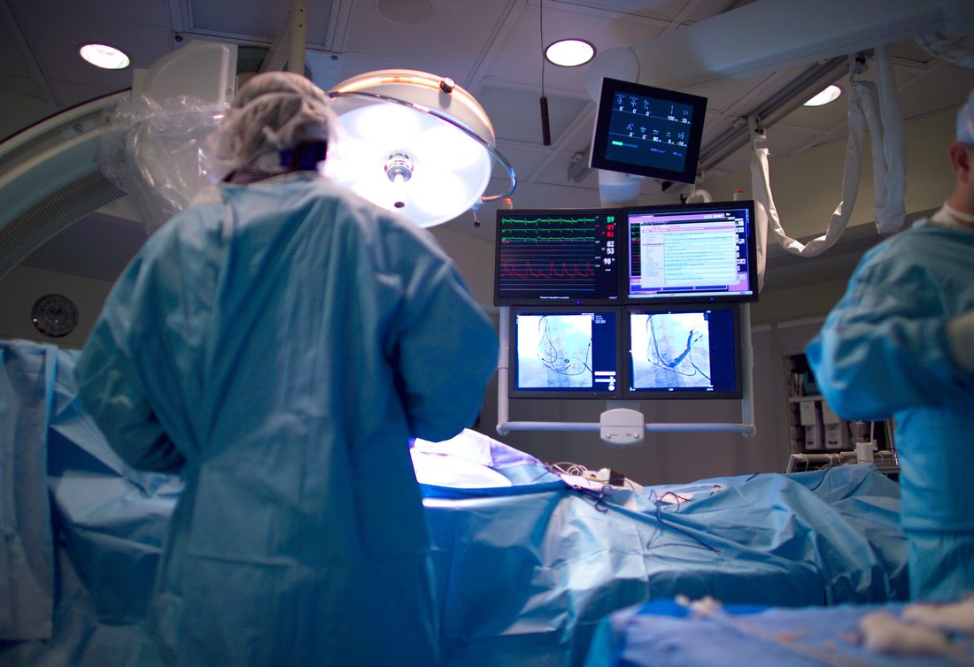 A team working under the lamp in a procedure room with vital signs and images running on the monitors above the table