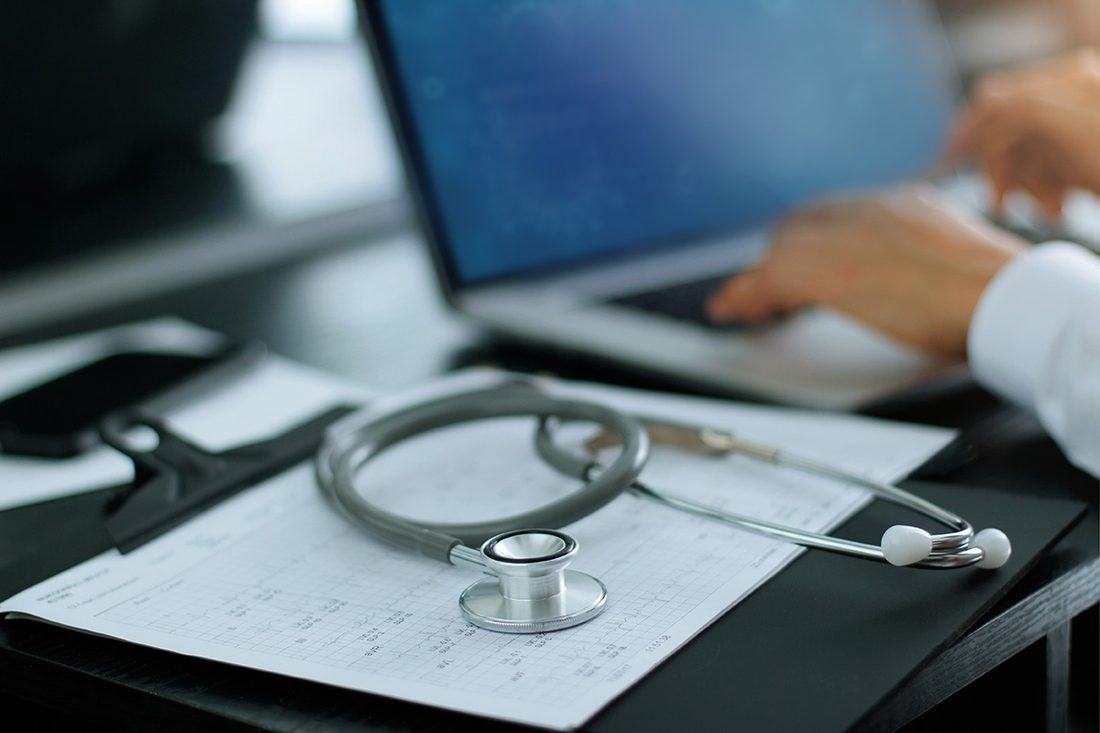 A clinician typing on a laptop beside a printed record with a stethoscope resting on it
