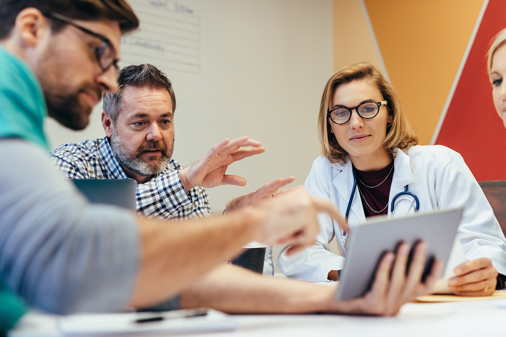 A clinician in a white coat and three colleagues around a table, reading something together on a tablet