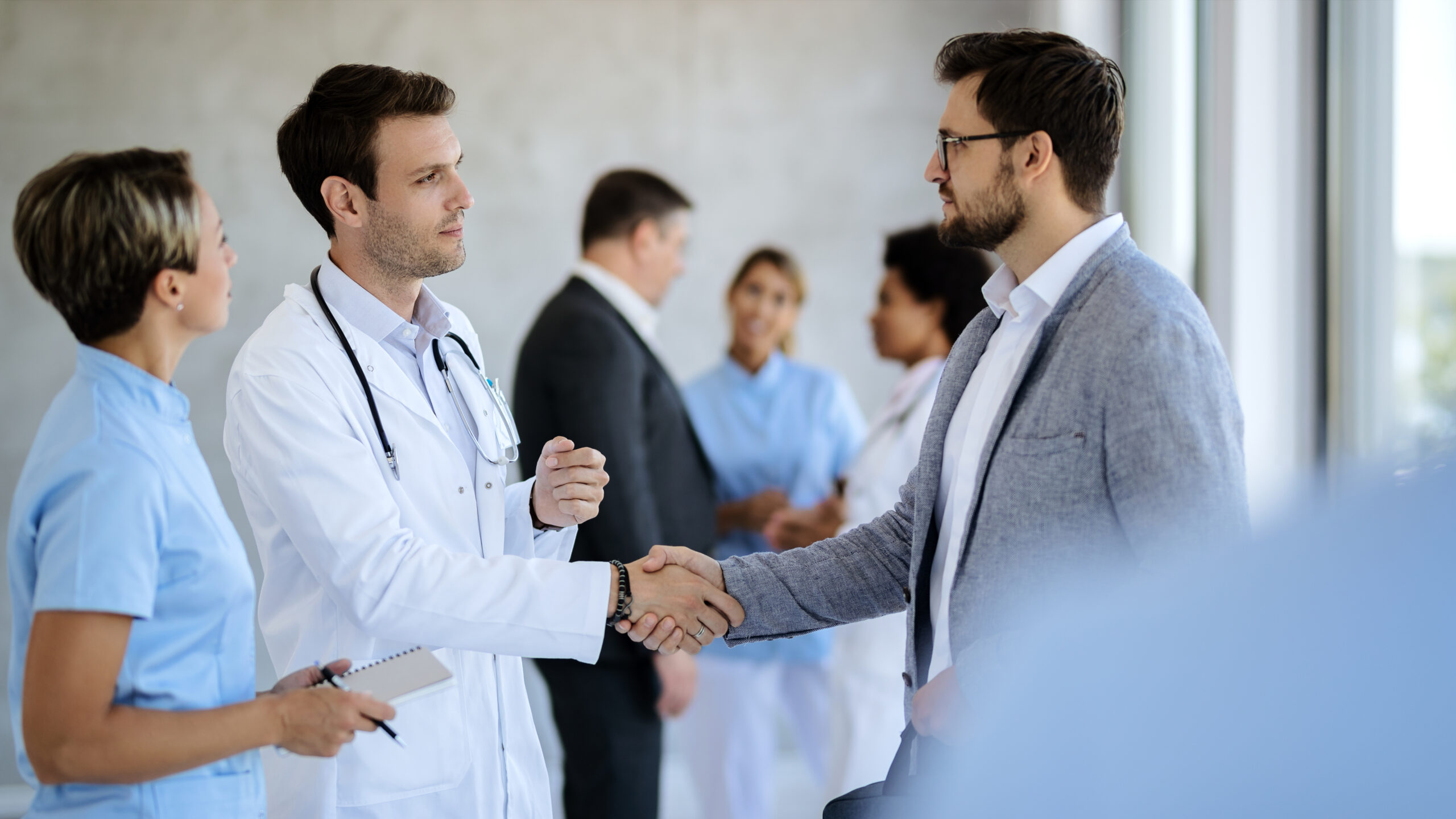 A clinician in a white coat shaking hands with a visitor in a jacket, colleagues talking in the corridor behind them