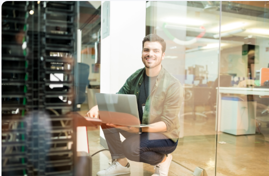An IT professional with a laptop crouching beside server racks in a glass-walled equipment room