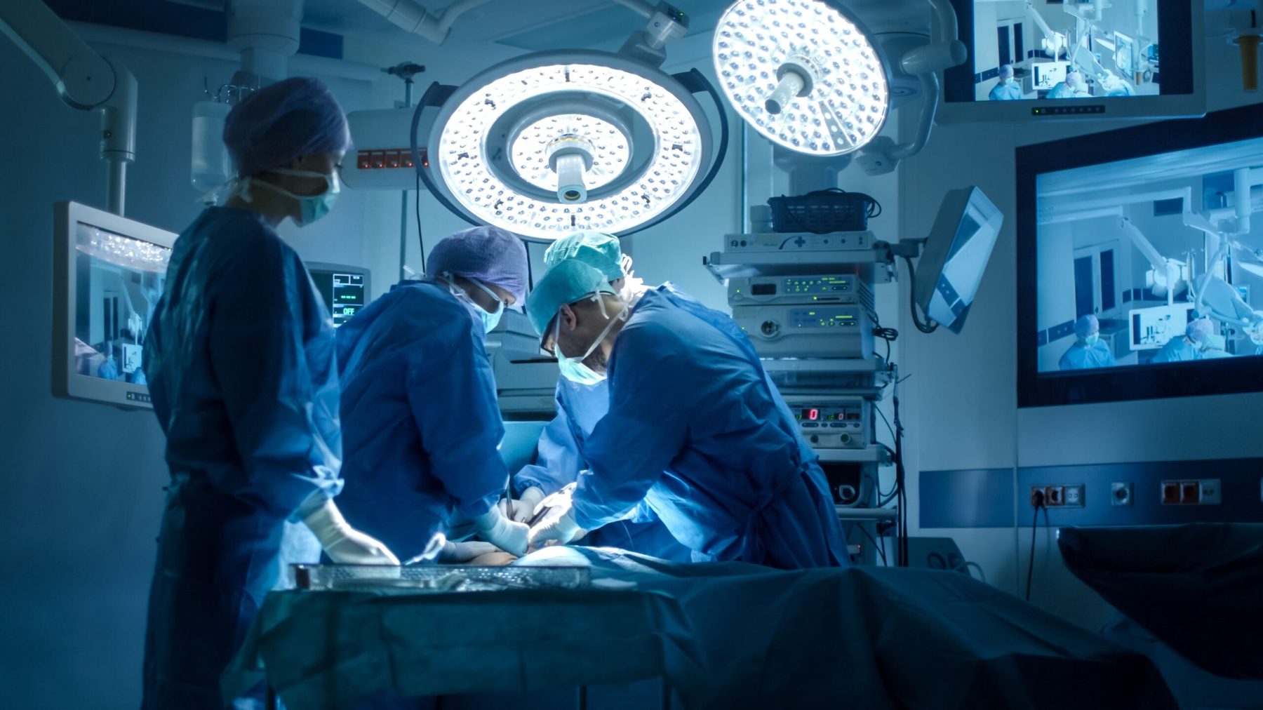 A surgical team at work in a darkened operating room while the procedure runs on wall-mounted displays